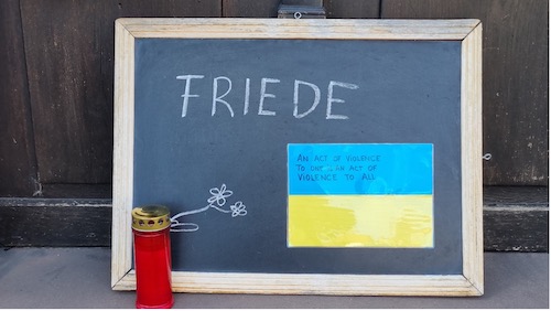 Ukrainische Fahne und das Wort Friede auf einer Tafel mit Grablicht davor